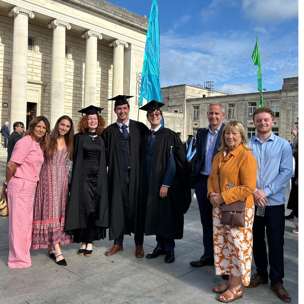 A group stands smiling outside the Guildhall, under a clear blue sky. In the center, three people in graduation gowns and caps with their family standing next to them.