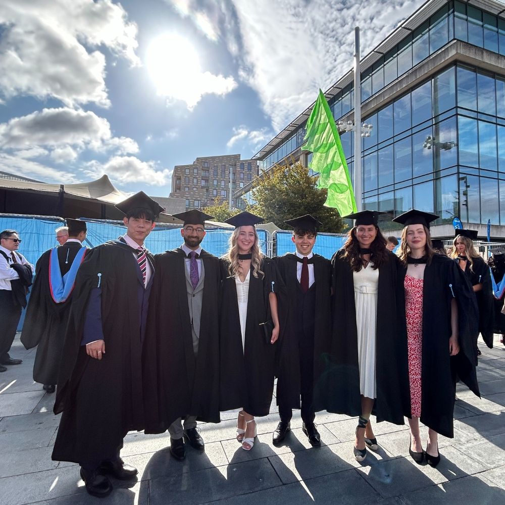 A group of six graduates in caps and gowns pose outdoors under a bright, partly cloudy sky. Behind them is a modern building with large glass windows.