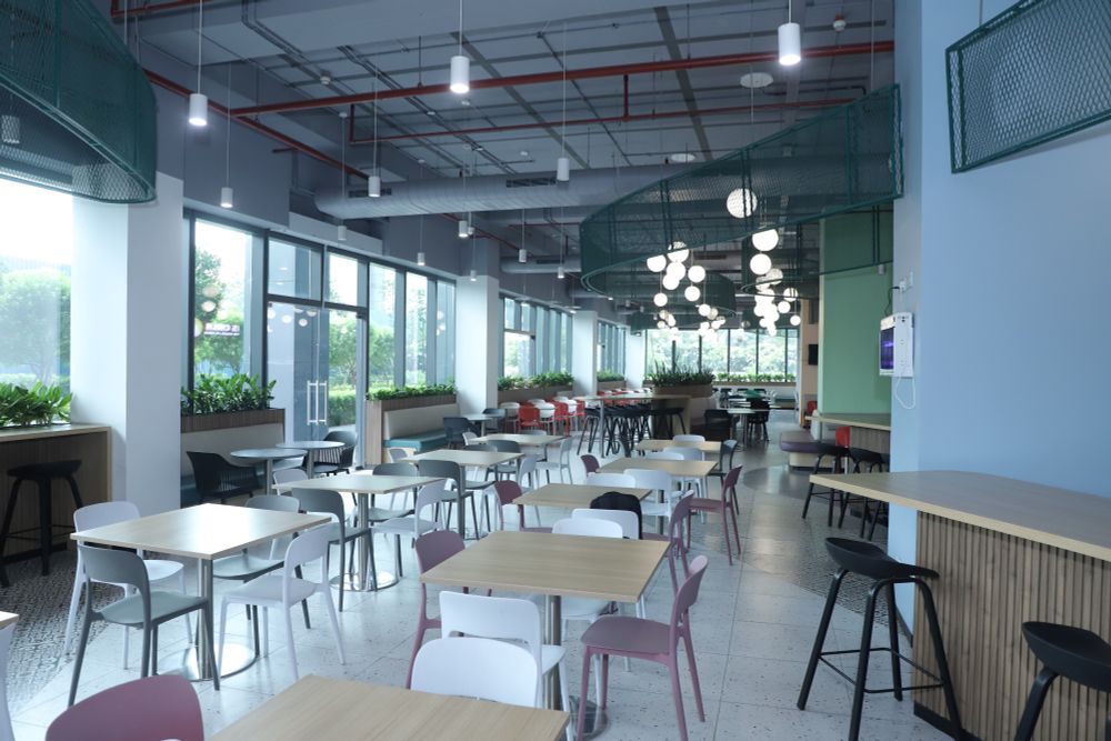 A spacious, modern cafeteria of University of Southampton Delhi campus featuring rows of square wooden tables with a mix of white, pink, and black chairs. 