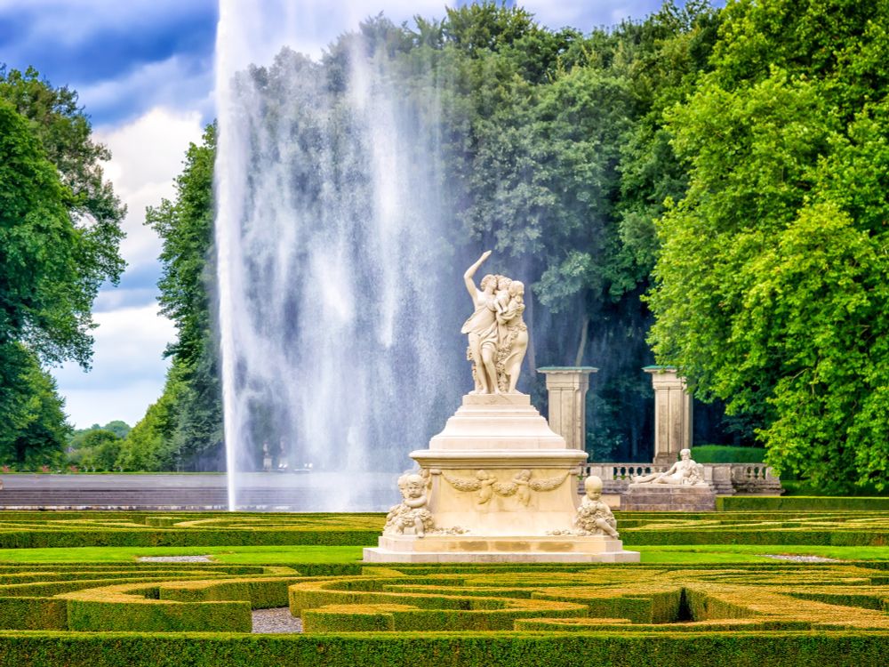 Parklandschaft mit Wasserfontäne, Säulen, geometrisch angelegten Wegen und einigen Bäumen.