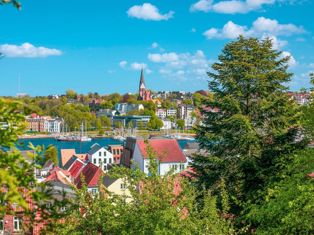 Blick auf eine Stadt mit Häusern und Bäumen und Sträuchern, in der Mitte ein Gewässer.
