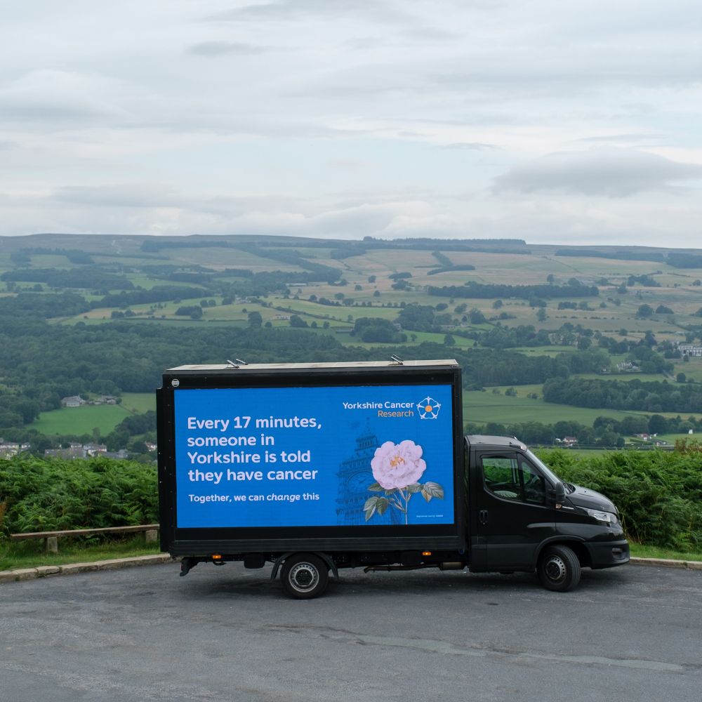 The Yorkshire Cancer Research digivan parked in front of scenic Yorkshire views of green fields and farmland. The wording on the van reads "Every 17 minutes, someone in Yorkshire is told they have cancer. Together, we can change this."