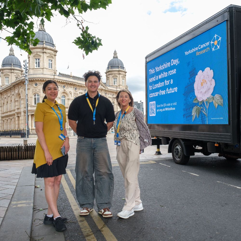 Three individuals wearing Yorkshire Cancer Research lanyards stand in front of a branded mobile advertising van calling for people to send a white rose to London for a cancer-free future. They are standing in Hull city centre, with historical architecture in the background.