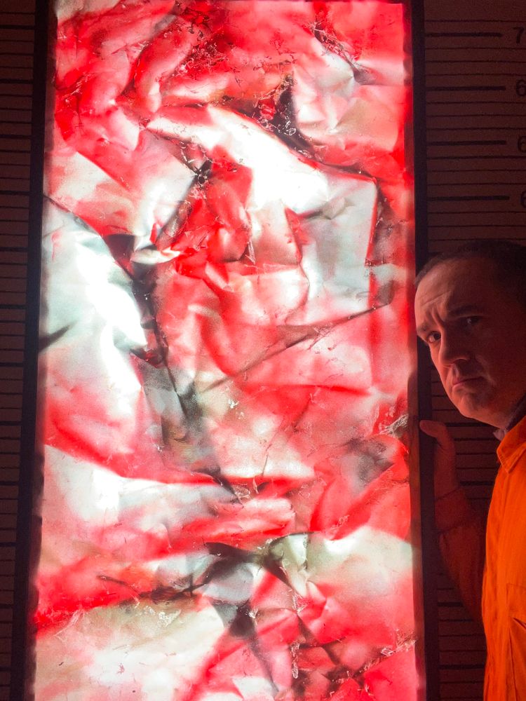 Obra abstracta “Dragontinas: Rubí” encendida desde dentro, mostrando tonos rojos, blancos y negros con pliegues que parecen llamaradas. A la derecha aparece el artista en penumbra, con expresión concentrada, mientras la luz interior de la obra resalta como un fuego que atraviesa la oscuridad.