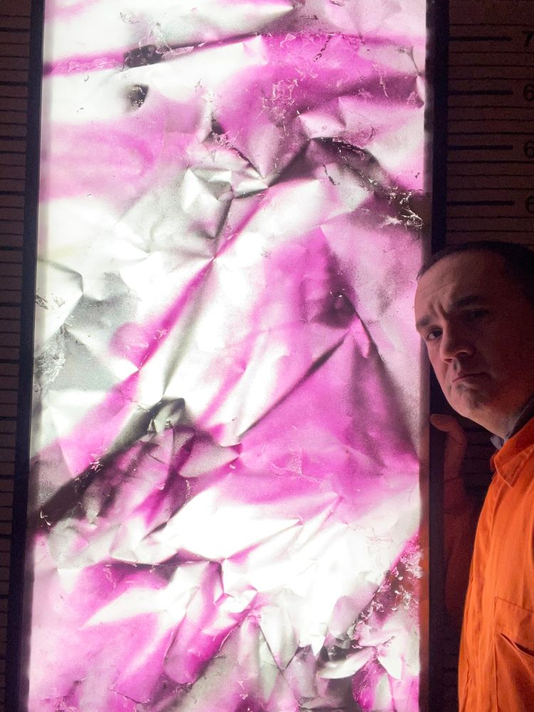 Obra abstracta retroiluminada de tonos amatista y blanco, con textura de vinilo arrugado que simula escamas de dragón. El artista aparece junto a la pieza encendida, evocando un portal de luz interior. Fotografía parte de la serie Dragontinas, arte contemporáneo inspirado en piedras preciosas y crecimiento personal.