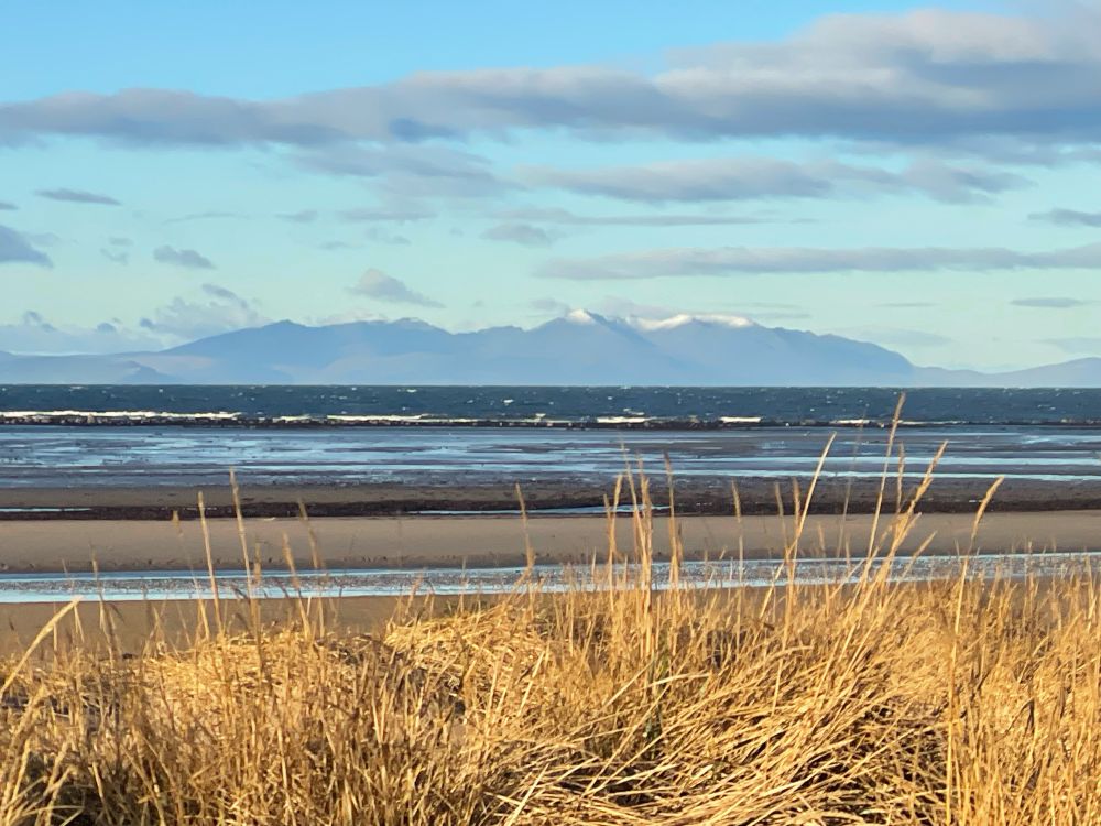 A photo of the Isle of Arran taken from the sand dunes in Ayr. There is snow on the high peaks of Goatfell and Caisteal Abhail. The sea has whitecaps, but the sky is mostly clear. 