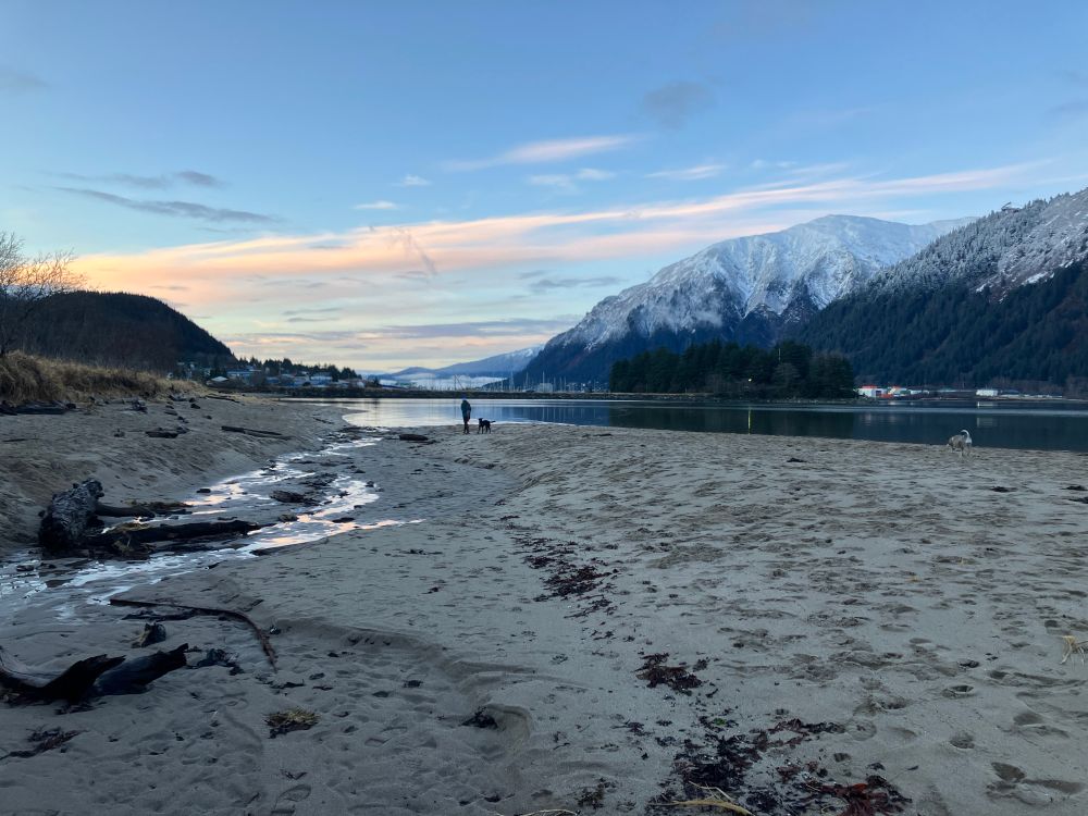 Beach in the late afternoon. A woman walking her dog in the distance. Termination dust in the mountains in the background.