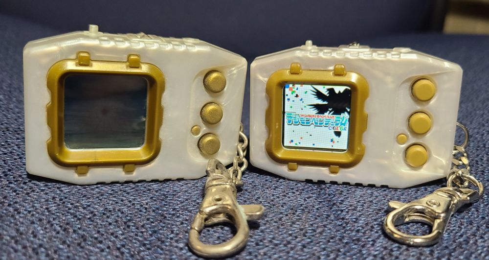 Big nerd sure did post some digimon virtual pets, oh lordy. They are pearly with gold borders and buttons and look almost entirely the same except the first is from 1998 and has a lcd screen and the second is less than a year old and has a color screen.