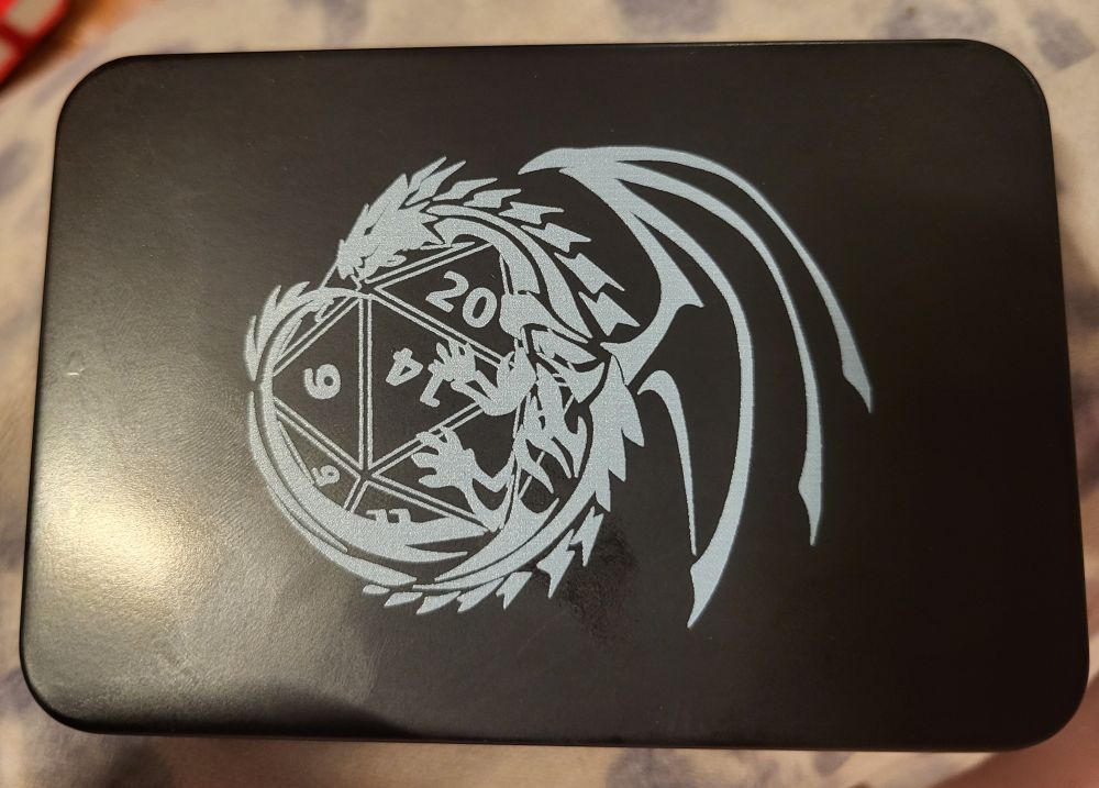 A DND dice tin, black, with a grey dragon on the lid, wrapped around a d20
