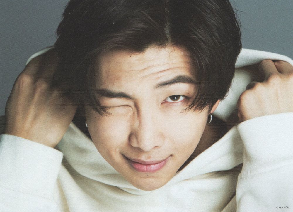 Kim Namjoon, aka RM of BTS, is staring into your soul while winking directly into the camera. The frame is close up on his shoulders and face. His left eye is open with left eyebrow arched in a wry expression. His black hair is long-ish, framing his face to just below his eyes. He's wearing a cream-colored hoodie & he is starting to pull up the hood. He has a killer lips closed smile and is seriously going to knock you over with the power of his stare. 💜💜💜