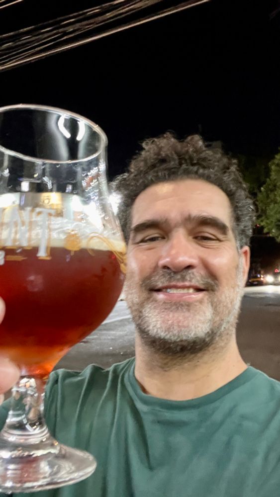 Eu, homem branco, cabelos castanhos, barba, camiseta verde, sorrindo e segurando um copo de cerveja bock.