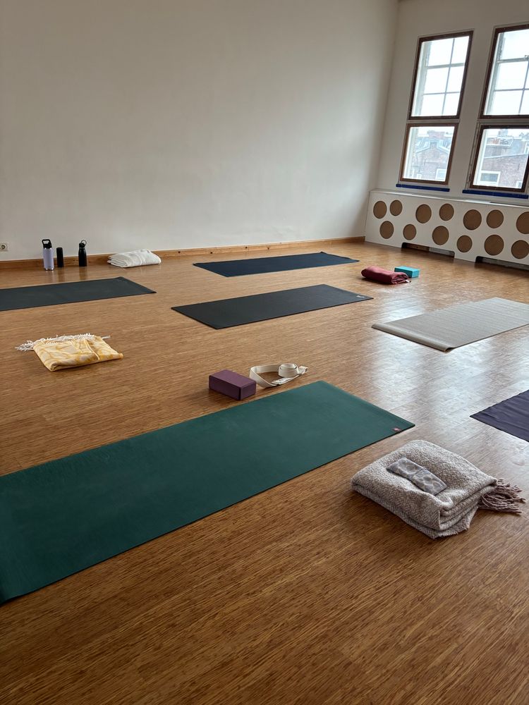 A room with a wooden floor and white walls filled with different colored yoga mats. There’s some windows letting though soft light. Blankets and yoga blocks are lying around to be used. 