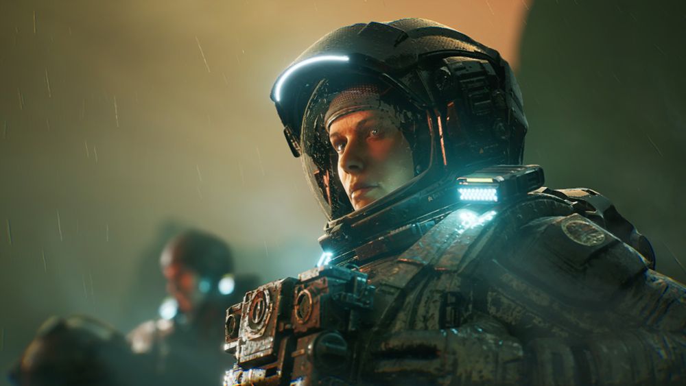 A close up of two astronauts, one near the camera and in focus, the other in the background and out of focus. The closer astronaut is illuminated by the lights of her suit and the red glow of the moon; the distant astronaut removes his helmet and examines it thoughtfully. Rain pours down, drenching them both.
