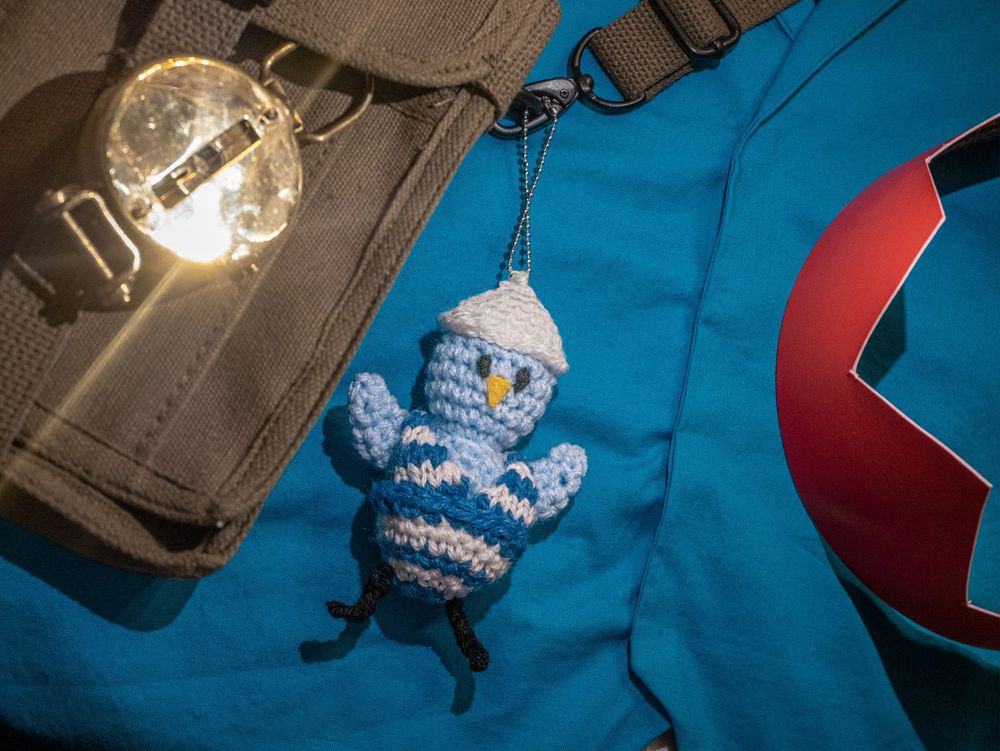 A close-up of a blue vest with no visible buttons, underneath an olive canvas satchel, from which hangs a shiny brass compass and a knitted plush of a blue bird in an antique bathing suit: SWIM BIRD. To the right, a familiar red paper crown.