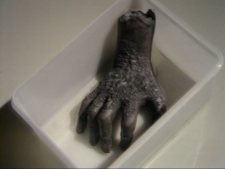 Screencapture of a gray, decrepit disembodied hand in a white plastic container