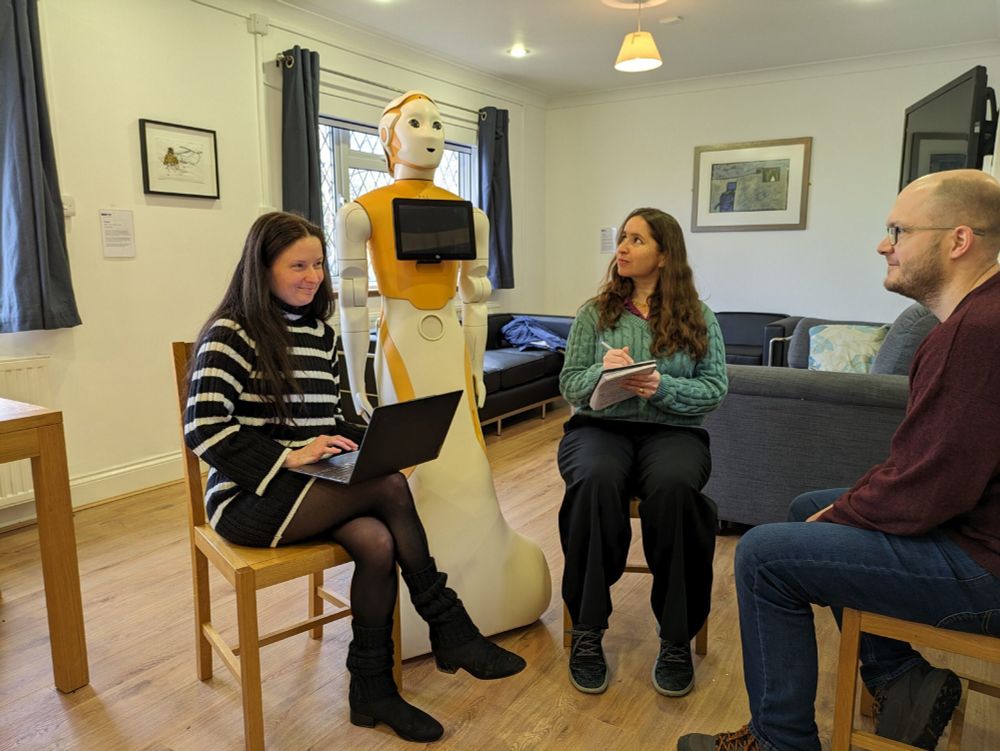 Three researchers sitting in a circle next to a large, white and yellow humanoid robot. Catherine is looking up at the robot and writing in a notebook.