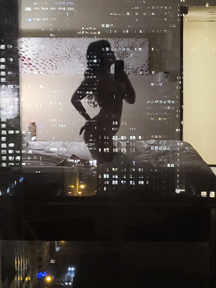 Ivy's reflection appears as a silhouette in a room with a view of the city. Also visible are the lights of San Francisco at night. 