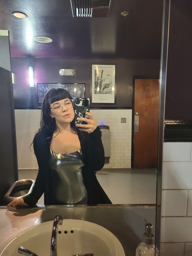 Ivy takes a mirror selfie. She wears a silver dress with a black cardigan.