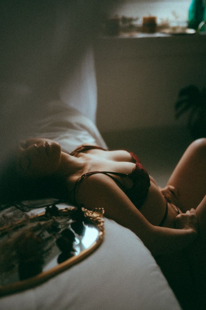 Ivy arches her back over a bed with her eyes closed. Soft light shines through a window. On the bed sits a mirrored platter covered in dried roses. Ivy wears a dark red lingerie set.