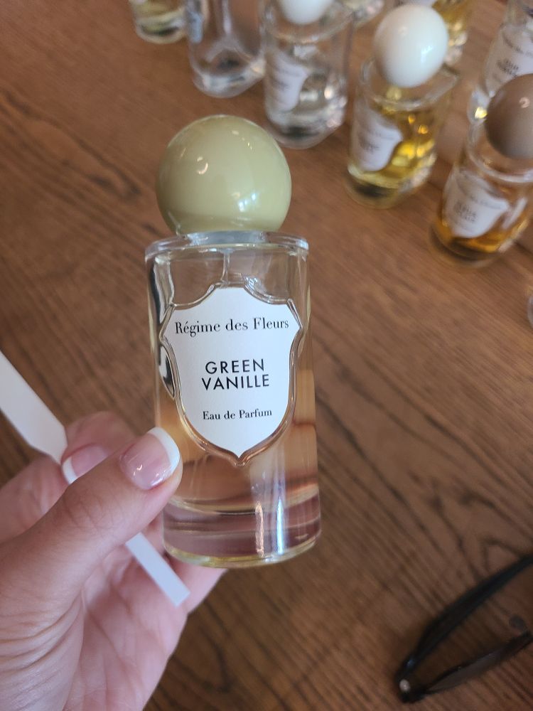 Ivy holds a perfume bottle in her freshly manicured, french-tipped hand. The perfume bottle is clear with a ball-like green cap. The bottle reads 
