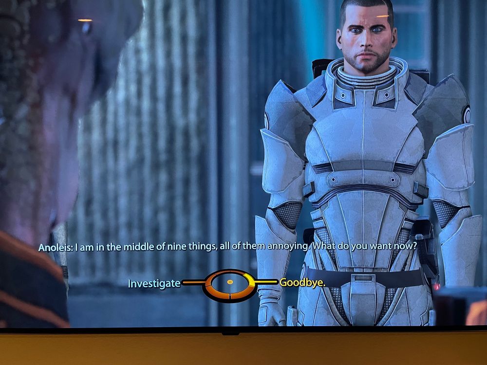Male Commander Shepard from Mass Effect wearing white armor. Dialogue on the bottom of the screen reads, “I am in the middle of nine things, all of them annoying. What do you want now?” 