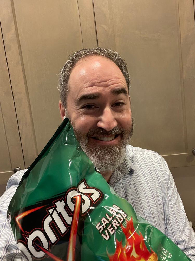 Jeff Akin smiling, wearing a button up blue shirt holding a bag of Salsa Verde Doritos. 