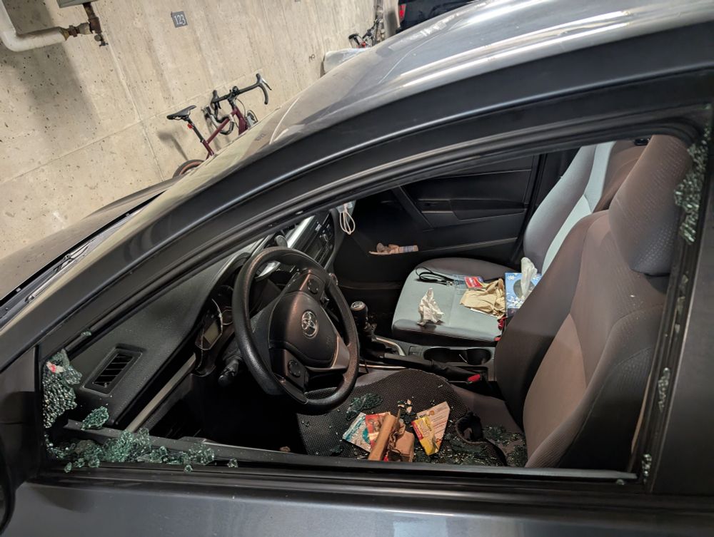 Upper part of a navy car's driver's side door showing a completely smashed window with some things on both seats (the things on the driver's seat are from the console in the door).
