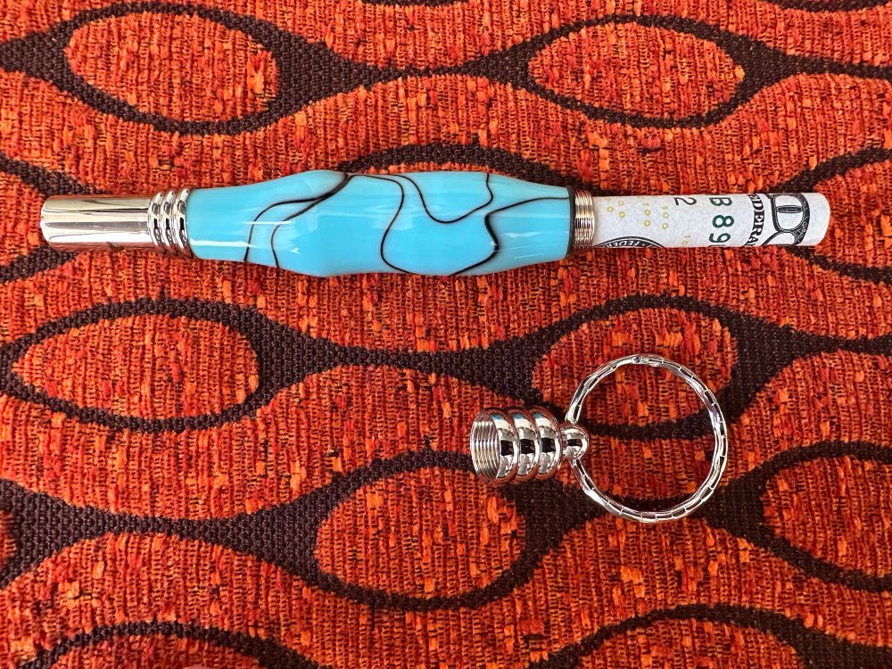 Handmade baby blue acrylic security whistle keychain with secret compartment. A $100 bill is rolled up sticking out of the keychain. Close up against orange and brown fabric backdrop.