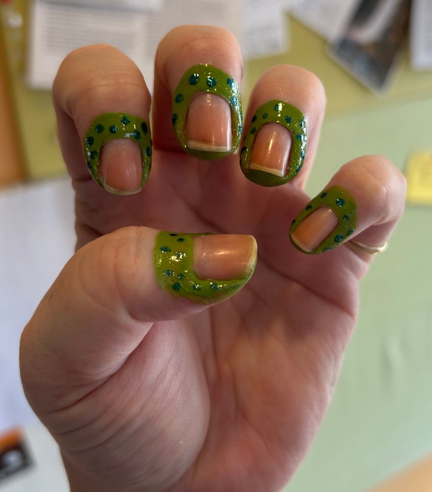 “Manicure” with skin painted around the nails but no polish on the nails. Skin is painted lime green and has sparkly blue dots all over it.