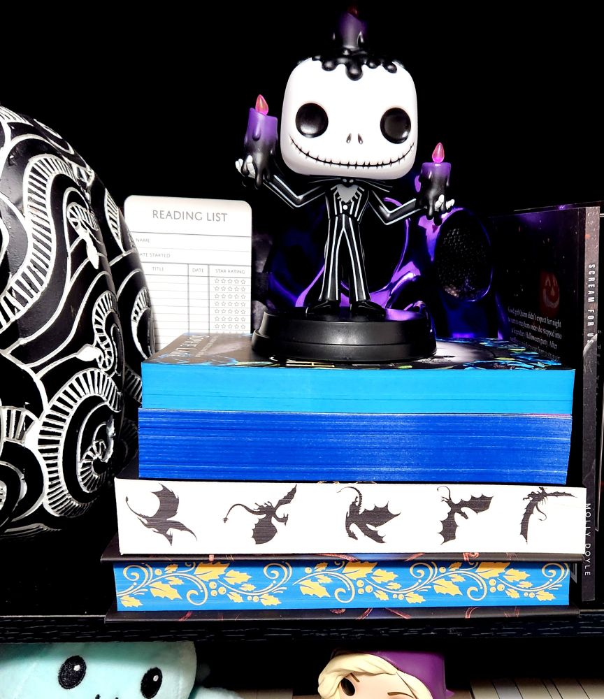 This picture is of my bookshelf, the shelves are a dark black and I have a small Jack skellington figure standing on top of books with sprayed edges, the Sprite edges very in hues of blue and holly, it also shows my fourth wing paperback with sprayed dragons. 