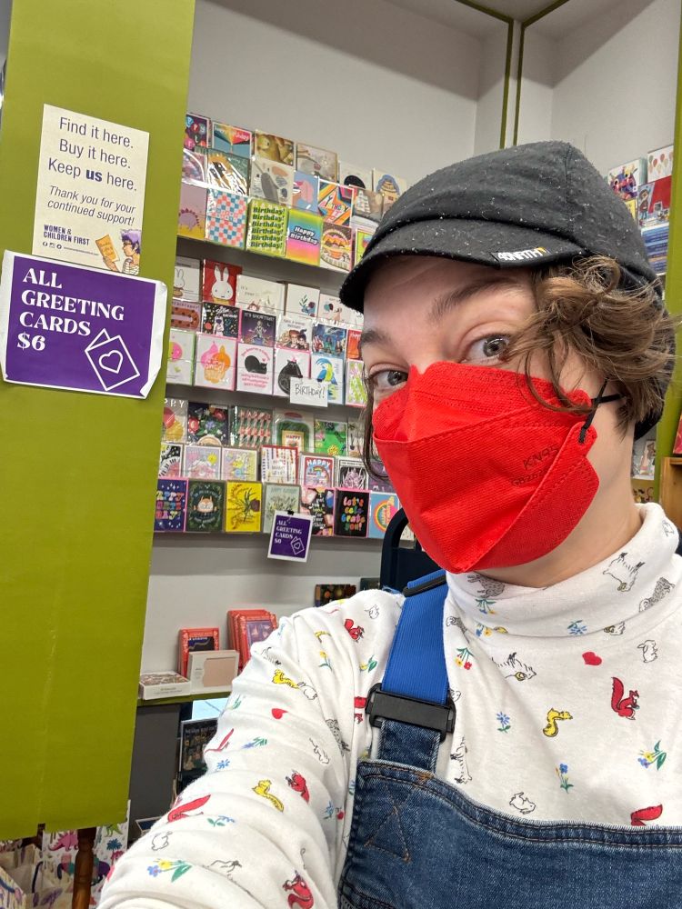 selfie in a red mask and black hat in front of the greeting card wall at women and children first