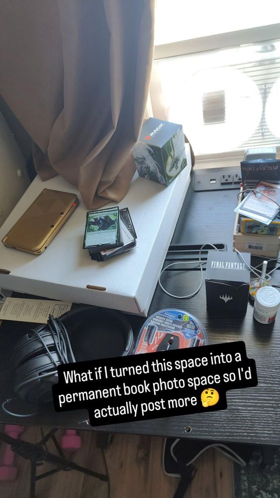 A photo of a desk next to a window and the desk is covered in magic cards and random items