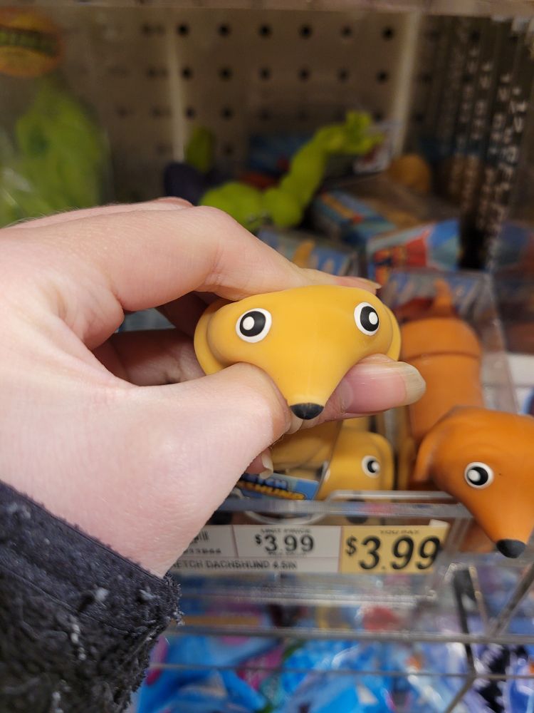Hand holding a squishy toy that looks like a brown Dachshund dog. Dog face is squished, so the muzzle is facing downward, and the eyes are facing the sides.