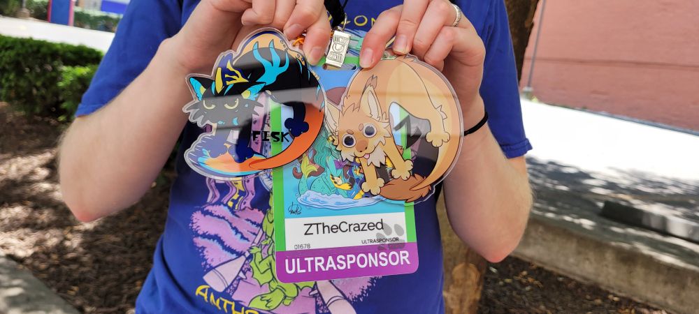 Image of a person's torso. They are holding 3 badges in their hand to show the camera. Two badges are of their furry characters, Fisk and Z. The third badge is their anthrocon registration badge with the text "ZTheCrazed" and "ULTRASPONSOR"