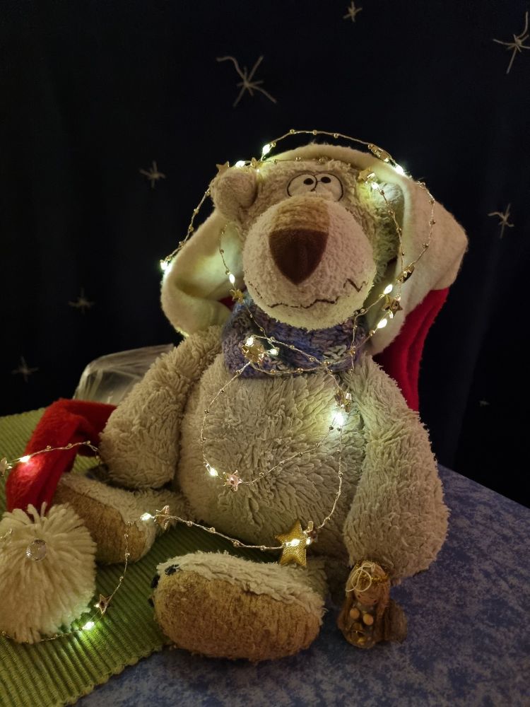 Ein weißer Bär mit Weihnachtsmütze auf dem Kopf, die sich hinter seinem Rücken nach unten schlängelt.
Eine leuchtende Lichterkette ist um den Kopf gewickelt und läuft in Wellen über den Bauch bis zu den Pfoten.