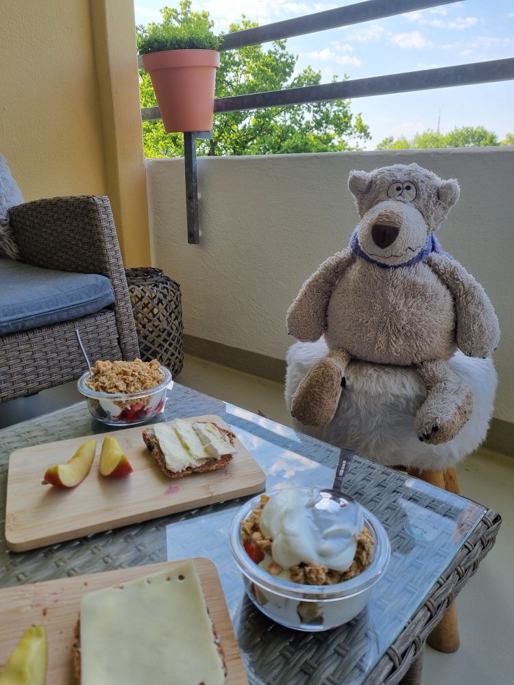Blick auf einen Balkon. 
Vorn im Bild ist ein Teil eines Balkones zu sehen, darauf stehen zwei kleine Schälchen mit Obst, Joghurt und Müsli sowie zwei Holzbrettchen mit Käsebrot sowie Apfelspalten.
Hinter dem Tisch sitzt ein weißer Bär auf einem Hocker, dessen runde Sitzfläche mit Fell bezogen ist.
