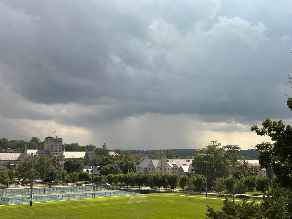A heavy rain shower in the distance, with buildings, tennis courts, and green grass in the foreground