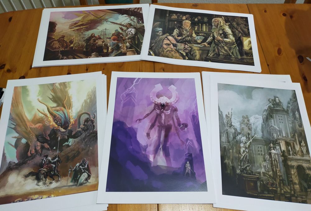 Five A3 prints arranged on a table, clockwise from top left: an image of several adventurers standing at a dockside dominated by a flying ship, a female knight seems to negotiate with a shopkeeper over a deck of cards, a cityscape made of tombs and mausoleums, a strange gigantic creature stalks a human warrior I'm hiding and finally a horse riding warrior rides to the aid of two warriors attacked by a dragon.