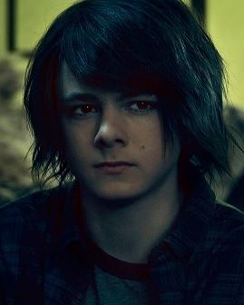 The Worst Minor in the World, Zach Goodweather from The Strain. A punchably-faced teen with shaggy hair and the eyes of a person with no self control looks out of frame, surely thinking about how else he can ensure the downfall of mankind on account of his refusal to use sense or think of anyone but himself. He is wearing a green and red or brown ringer shirt and a darker collared shirt over it. His face says "vampire mommy says I'm a good boy and that everyone else is bad". 