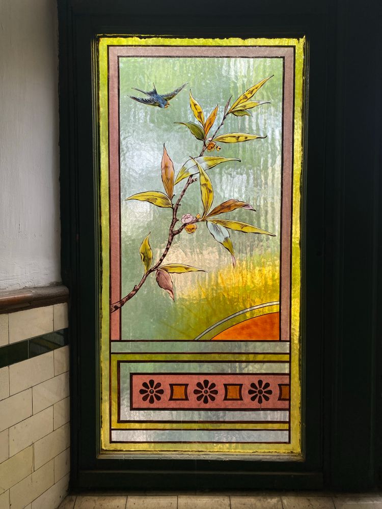 This is an image of a stained glass panel. The panel features a bird in flight and a branch with floral detail. 