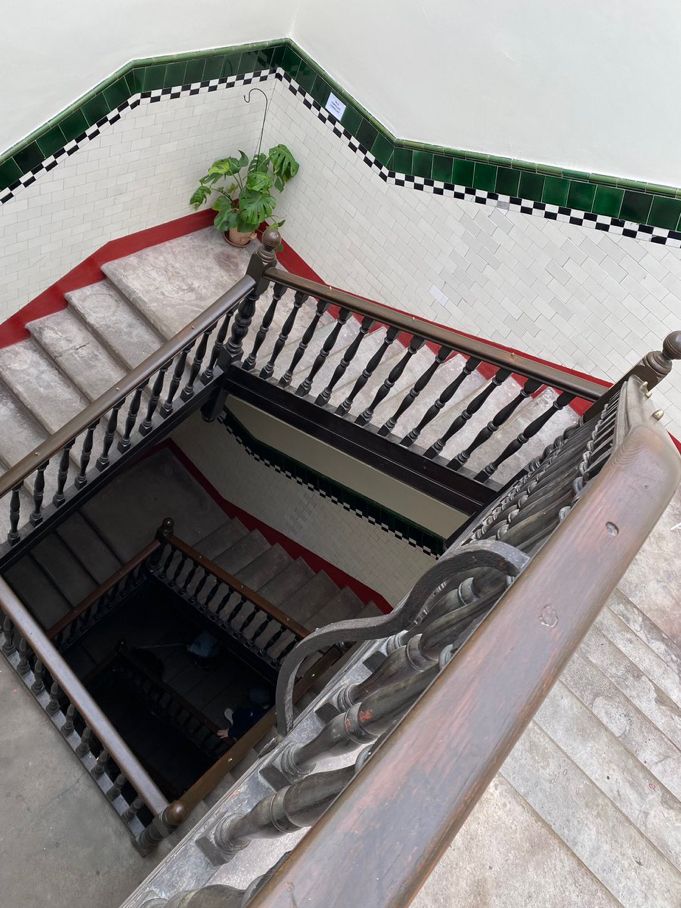View of a three floor stairwell. The staircase is squared off, allowing a shadowy view down to the ground floor. The walls are lined with chequered subway style tiles. The steps are a cool grey concrete with visible wear. There is a small green plant on the corner step. 