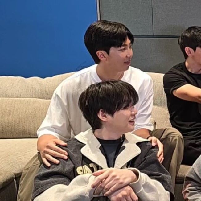 yoongi and namjoon from BTS. they are both looking to the left. namjoon is sitting on a couch with his legs spread and yoongi sits on the floor in front of namjoon and is framed by his knees. namjoon's hands rest on yoongi's shoulders. they look happy and comfortable.