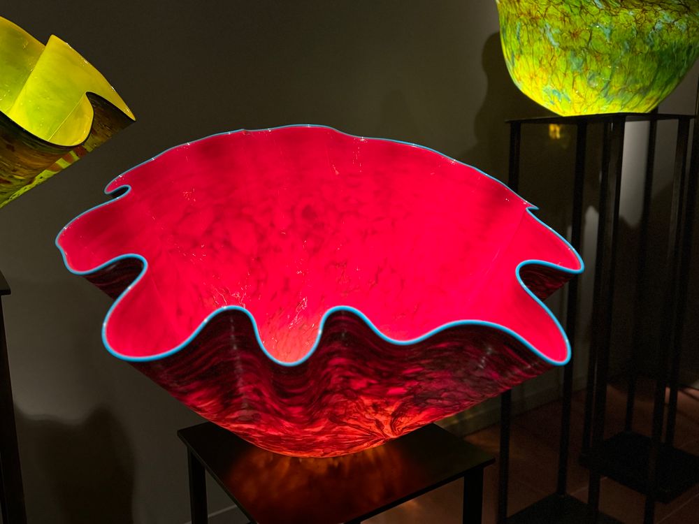A large translucent red glass bowl with a rippled edge lined with white glowing from an unseen light source. There are several other bowls in the darkened room of various colors.