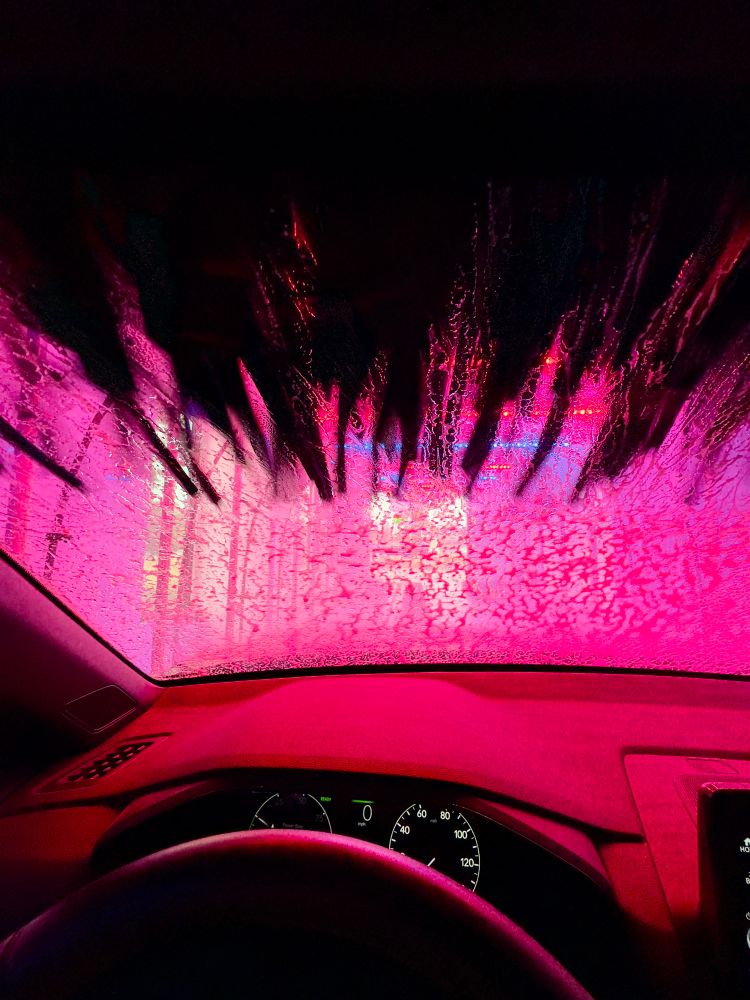 Colorful red lights shine upon a car dash as the automated car wash brush comes down upon a soap covered windshield.