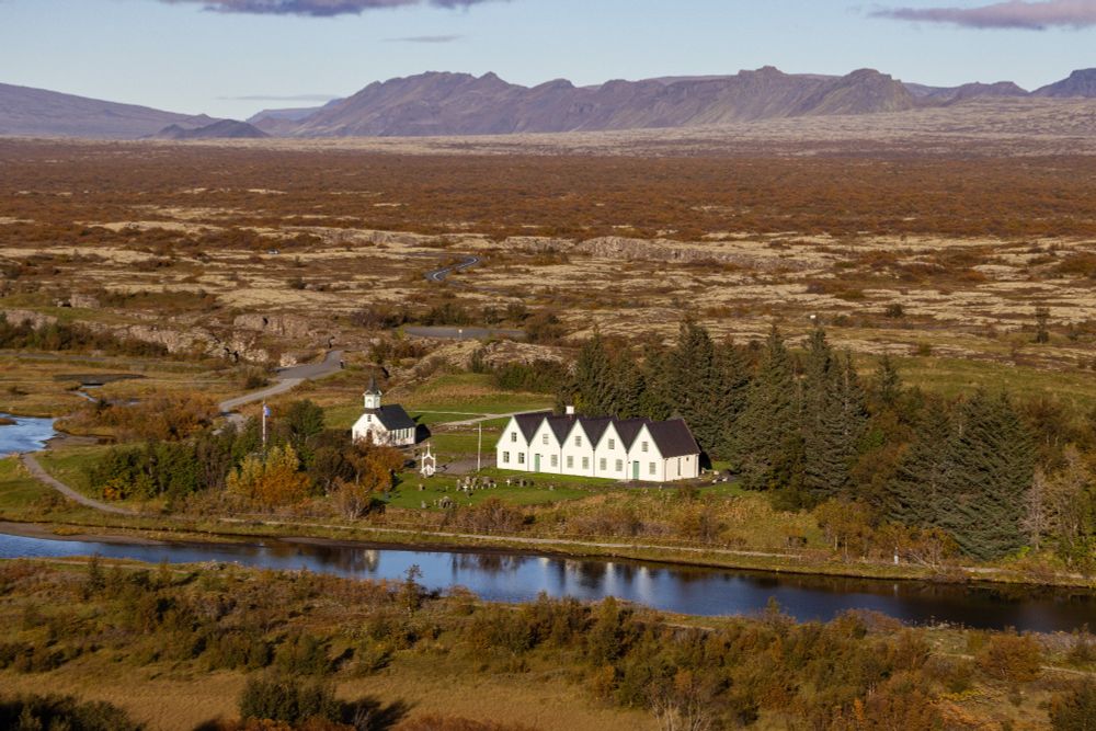 Das Bild zeigt eine ruhige, weitläufige Landschaft. Im Vordergrund verläuft ein schmaler, gewundener Fluss mit klarem Wasser, der sich durch eine grüne Ebene schlängelt. Entlang des Flusses stehen einige weiße Gebäude mit dunklen Dächern, darunter  eine kleine Kirche und angrenzende Häuser im traditionellen isländischen Stil. Diese Gebäude sind von Bäumen umgeben, die sich deutlich von der ansonsten kargen Vegetation abheben. Die Umgebung ist geprägt von felsigem Terrain mit spärlichem Bewuchs, das sich bis zu den entfernten Bergen am Horizont erstreckt. Die Berge wirken dunkel und massiv, und darüber spannt sich ein klarer, blauer Himmel ohne sichtbare Wolken. Eine schmale Straße führt durch die Landschaft zu den Gebäuden und unterstreicht die Abgeschiedenheit des Ortes.