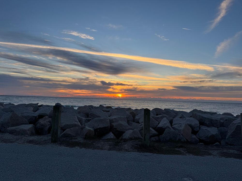 The sun rises in a strip of orange cutting through a swath of gray clouds over the ocean 