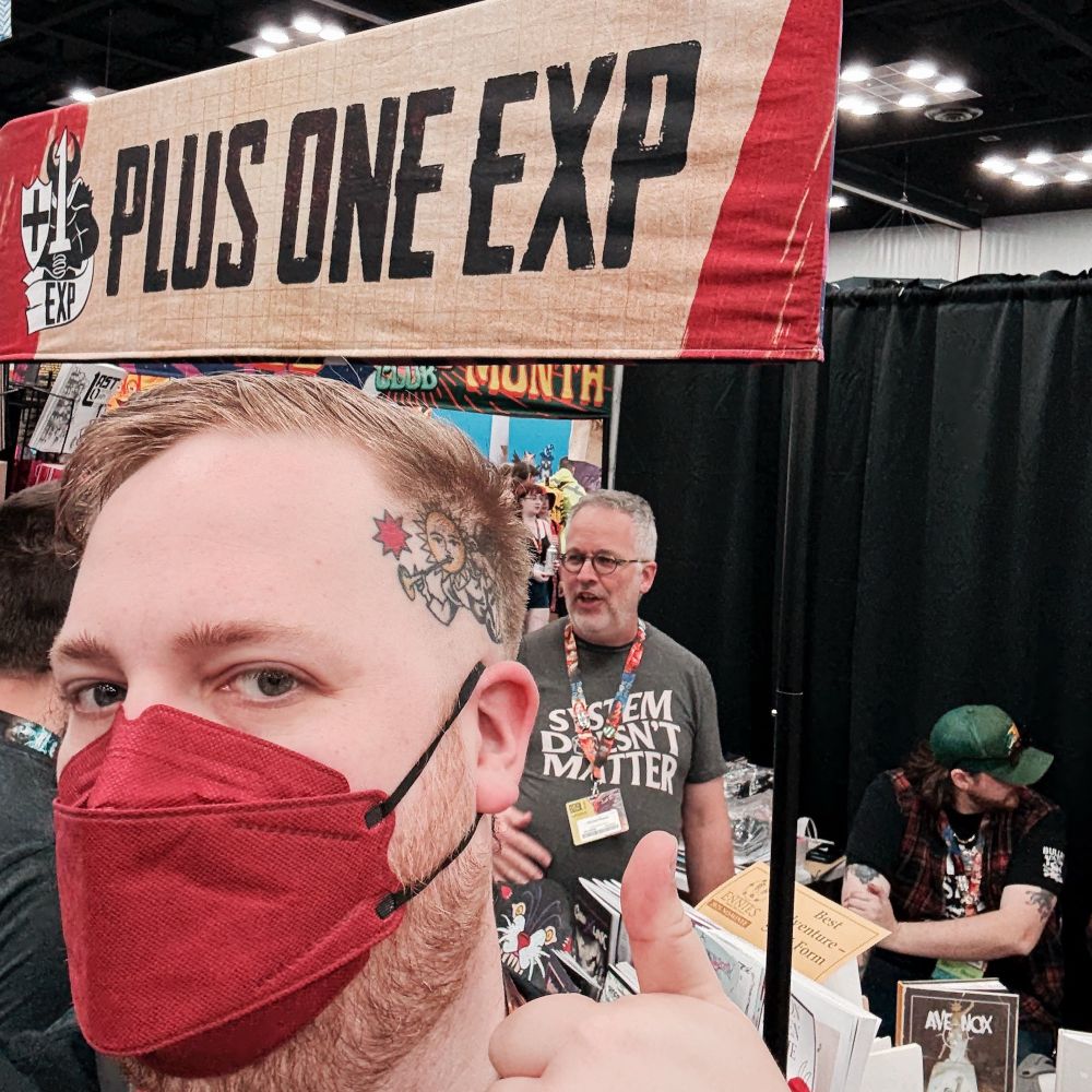 A white person with head tattoos and a red facemask giving a thumbs up in front of a convention booth banner that says plus one exp 