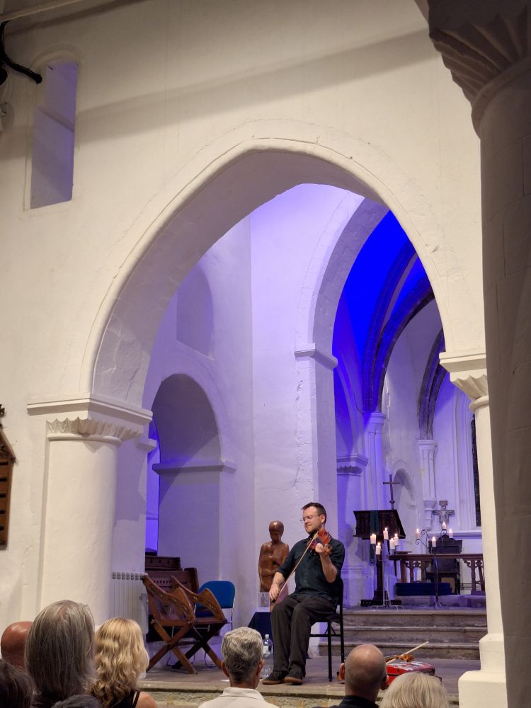 A solo fiddle player, seated, in a building with tall, white arches and a high ceiling with some strong blue shadows above and behind him. It's Sam Sweeney playing at a church in Guildford, St Mary's. 