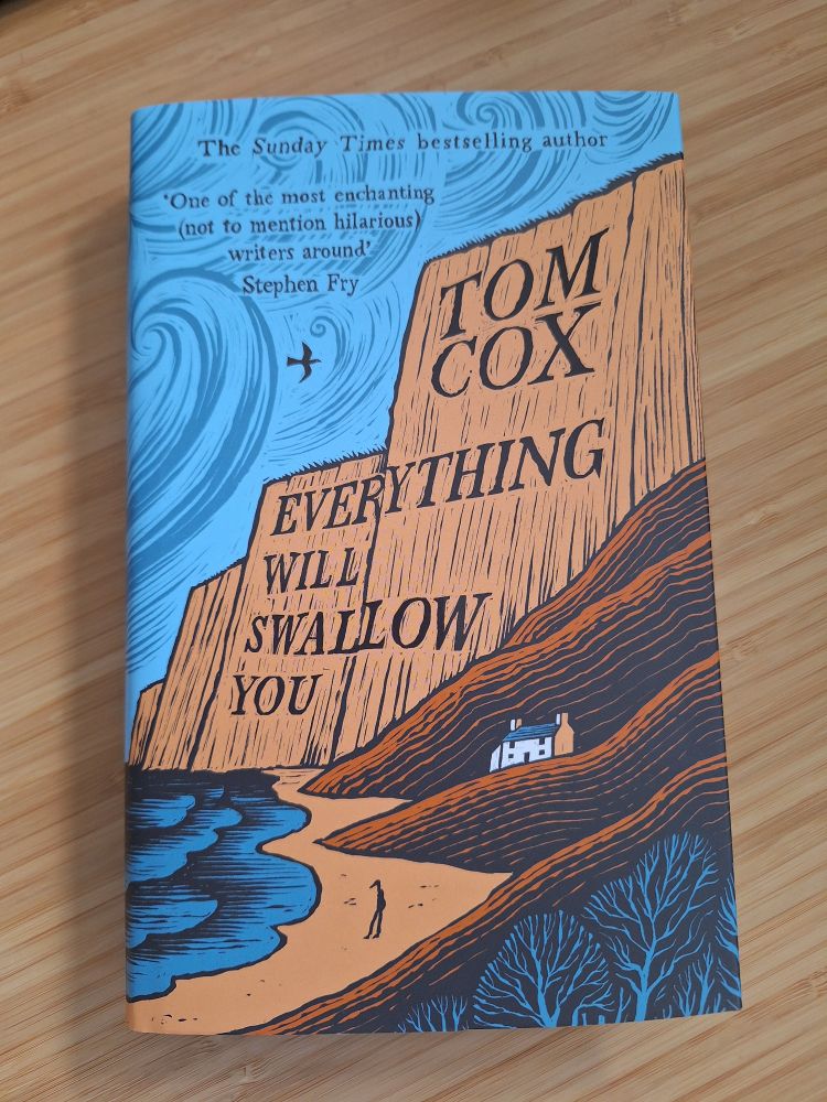 A hardback book, on a wooden desk. It's the newly published "Everything will swallow you" by Tom Cox. Suitably stylised beach/sea/cliffs cover design with figure, house, trees and a gull.
