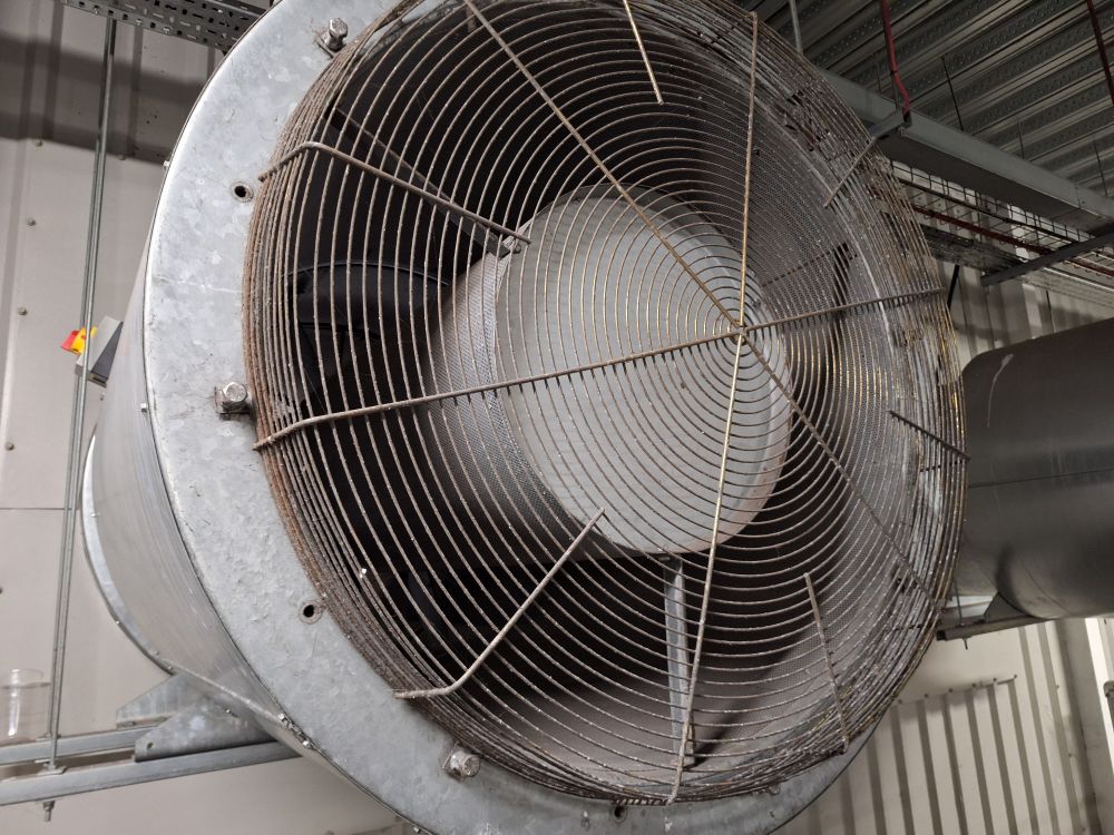 A large industrial fan in a building, galvanized grey cowl, mesh etc. Second one visible to its right, the scene being shades of pale silvery grey colours apart from a tiny dot of yellow and red from the power switch upper left.